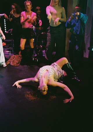 Bildet viser en person som ligger dramatisk bakoverlent på gulvet i en sceneopptreden, iført et mønstret antrekk og høye støvletter. Rundt står en gruppe i ulike positurer, hvor noen peker, klapper eller fotograferer henne, mens stemningen er opplyst av fargerikt scenelys som skaper en intens og energisk atmosfære.