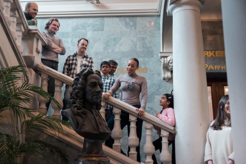 Gruppe med barn og voksne står i trapp med marmorrekkverk og byste i forgrunnen, omviser forklarer.