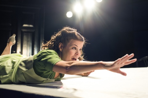 Kvinne i 40-tallskjole og -frisyre ligger på magen på et bord, gaper og strekker hånda frem