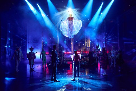 En person i stor kjole henger over en scene fylt av blått lys. På gulvet står folk og ser opp mot henne