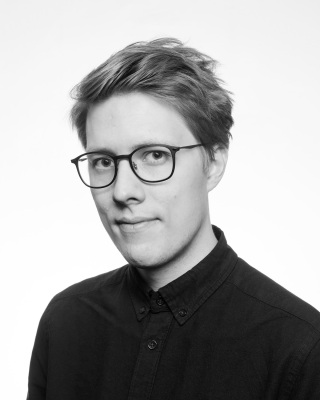 Svart-hvitt-bilde av mann i 20-/30-årene med briller og kort hår iført skjorte. Han er skrått vendt mot, og ser inn i, kamera