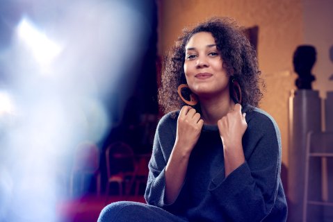Kvinne i 30-årsalderen i blå genser og store runde øredobber sitter og ser smilende lurt inn i kamera. Hun holder i kragen med begge hender. I bakgrunnen skimter vi gul vegg og en byste.