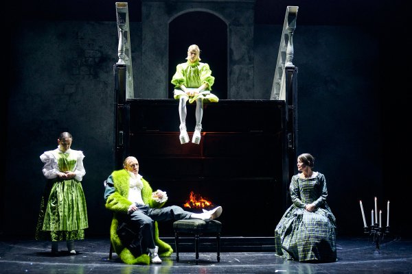 Fire skuespillere i store 1800-tallsinspirerte fargerike kostymer sitter rundt en peis