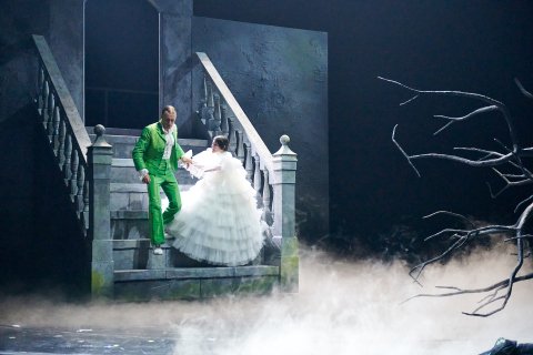 Mann i grønn dress og kvinne i stor brudekjole går ned en trapp på en mørk og røykfull scene