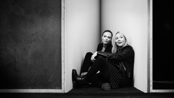 Svart hvitt bilde av to kvinner i mørke klær, som sitter på gulvet trykket sammen i et hjørne. De ser hver sin vei med et lite smil.