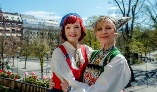 To kvinner i bunad holder rundt hverandre og smiler. De står ute på blomstrete balkong