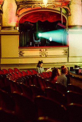 To jenter og en kvinne sitter i teatersal. Kvinnen leser i et manus