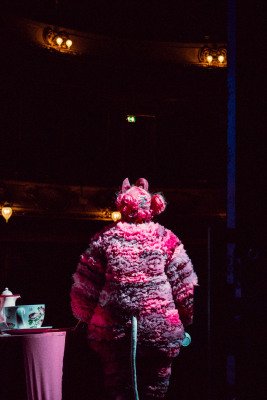 En skuespiller i digert rosa musekostyme står med ryggen til ved et bord med en diger tekopp