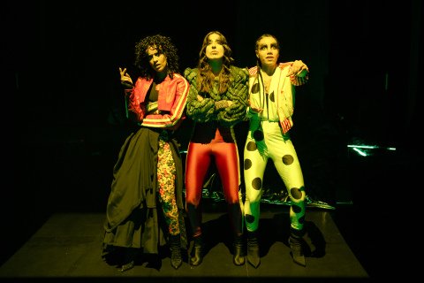 Tre kvinner i fargesterke klær på en mørk scene