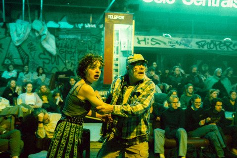 En ung kvinne og eldre mann står foran en rød, gammeldags telefonkiosk, omgitt av publikummere.