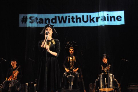 Fire kvinner på en scene, en foran med mikrofon, tre bak med instrumenter, iført svarte folkedraktaktige kostymer. Bak henger et banner med #standwithukraine