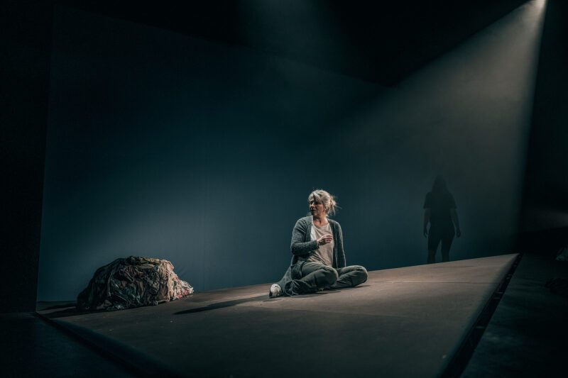 Kvinne sitter på mørk scene, i bakgrunnen står en annen kvinne