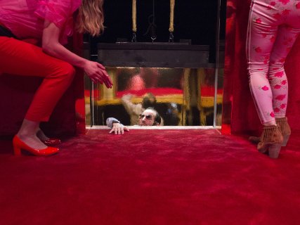 Mann står i en heis som har satt seg fast mellom etasjer. To kvinner i høye hæler står i en rød lobby og prøver å hjelpe ham