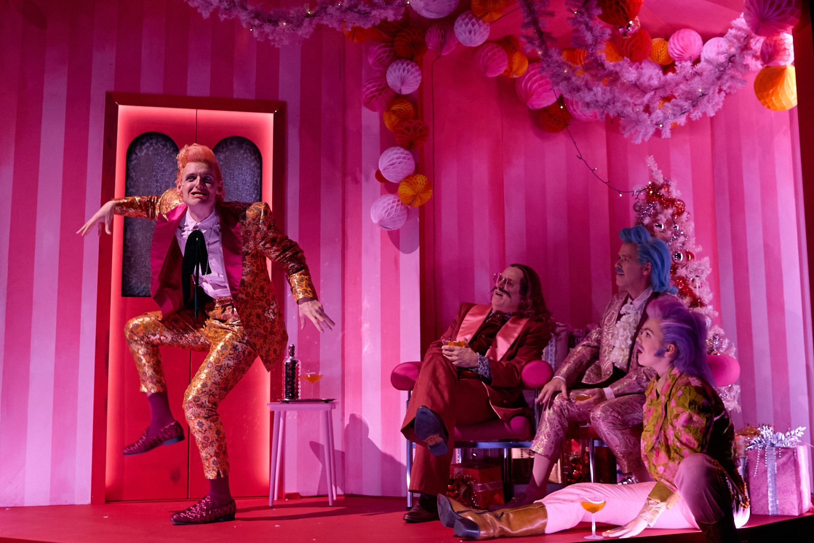 En karakter med oransje og rosa dress står i en energisk positur i et rom med rosa stripete vegger. Rommet er pyntet til fest, med en hvit juletre og dekorasjoner. Tre andre karakterer med fargerike klær sitter og ser på.