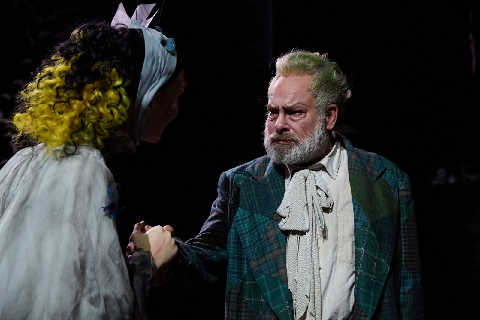 En scene der en eldre mann med grått skjegg og grønt hår, iført en rutete dress og hvit sløyfe, holder hendene til en kvinne med og gule krøller. Mannen gråter, og de står nært hverandre i et dramatisk lys.