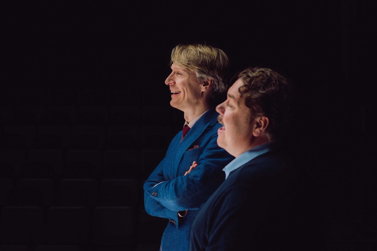 To menn står ved siden av hverandre, smilende og ser ut til høyre for kameraet. Den ene har på en blå dressjakke, den andre en mørk genser med en blå skjorte under