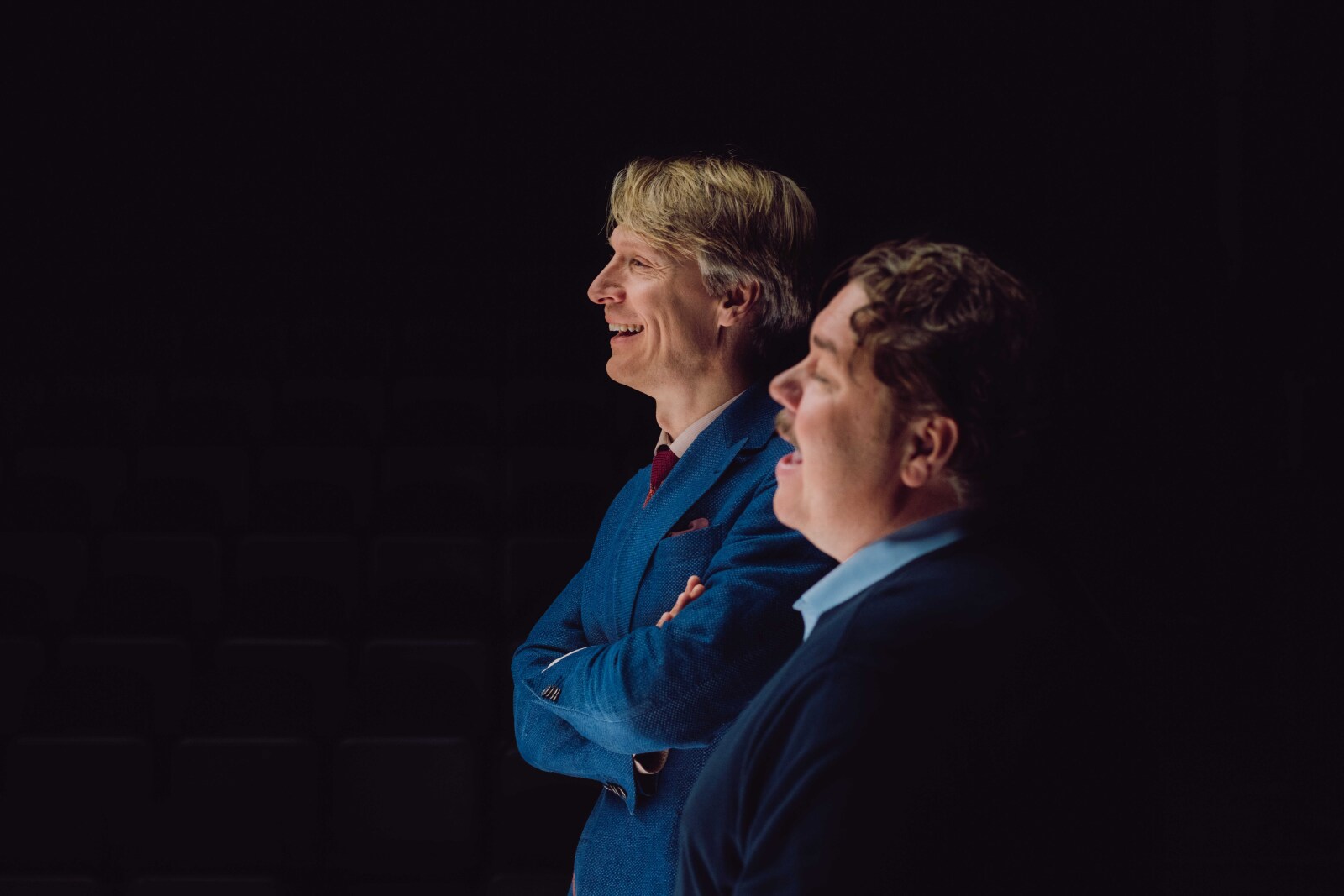 To menn står ved siden av hverandre, smilende og ser ut til høyre for kameraet. Den ene har på en blå dressjakke, den andre en mørk genser med en blå skjorte under