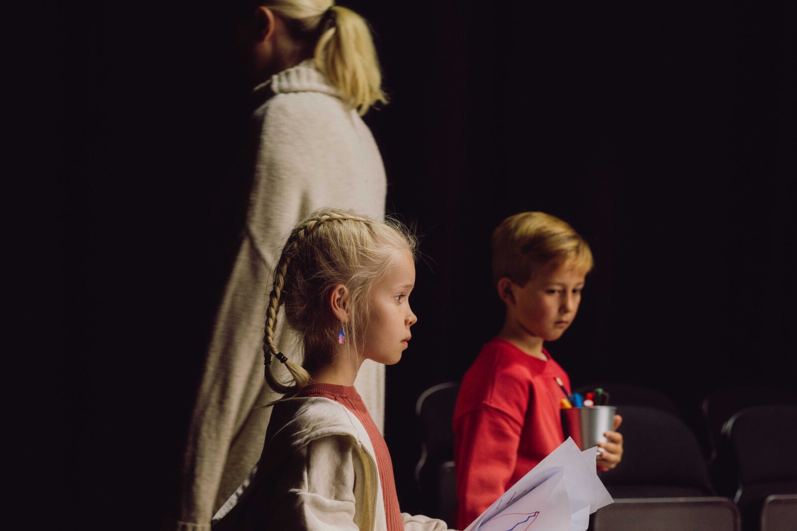 En ung jente med fletter ser konsentrert ut mens hun holder tegninger. En gutt med lyst hår står i bakgrunnen, holder en kopp med tusjer, og en kvinne med lyst hår, sett bakfra, er på vei bort.