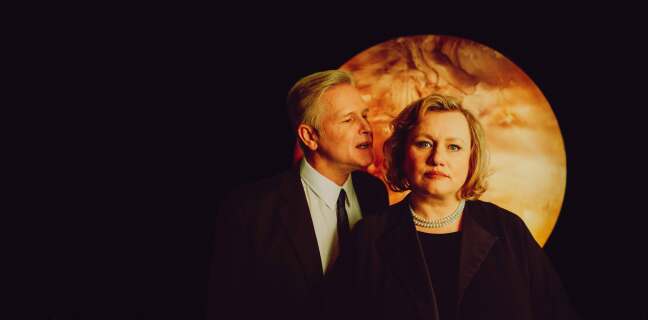 Mann kjefter på kvinne. Hun ser i kamera. De står penkledd foran en måne