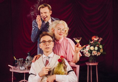 to menn og en kvinne i feststemning som står bak hverandre og drikker champagne og røyker. Mannen i front sitter med en høne i fanget.