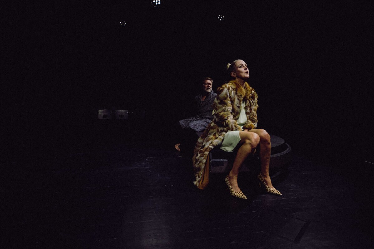En person i pelsjakke og leopardmønstrede sko sitter på en benk i en dramatisk, mørk belysning med en annen person i bakgrunnen.