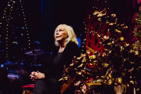 En kvinne med lyst hår sitter på en stol. Hun er kledd i svart og er omgitt av julepynt og dekorasjoner med gulltema.