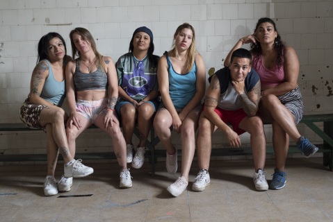 Seks kule lettkledde personer sitter på en benk i det som ser ut som en fengselscelle