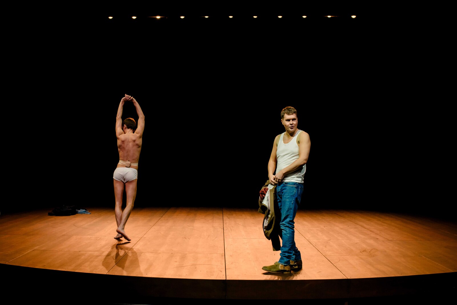 En mann i hvit singlet og jeans står på scenen med klær i hånden, mens en annen mann i hvite undertøy strekker armene over hodet med ryggen til.
