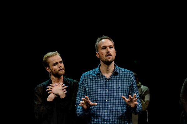 To skuespillere på scenen – En person med skjegg og sort antrekk holder hendene mot brystet, mens en annen i rutete skjorte står ved siden av og gestikulerer.