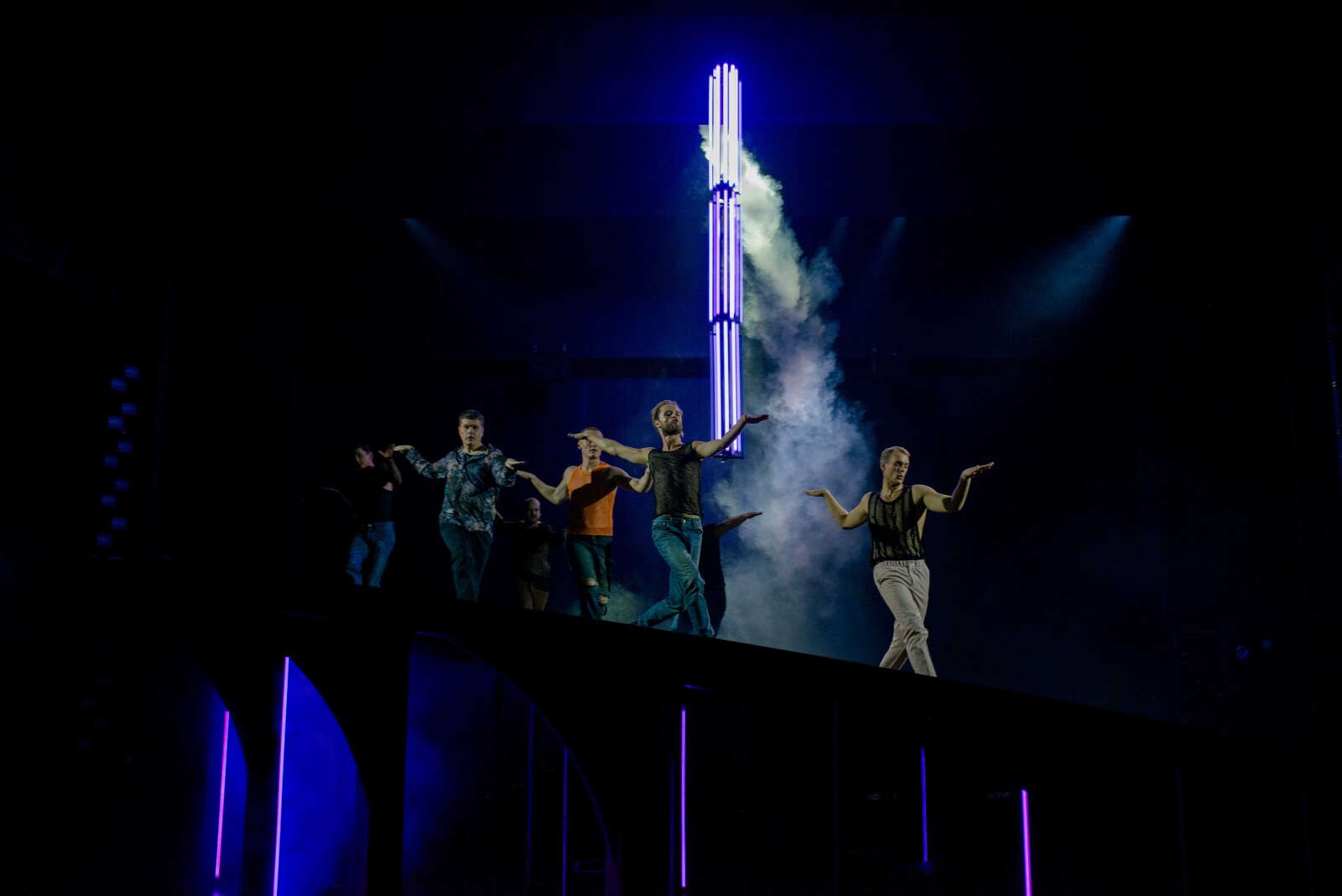 En gruppe menn danser i formasjon på en opplyst scene med blått lys og røyk, med vertikale lysrør i bakgrunnen.