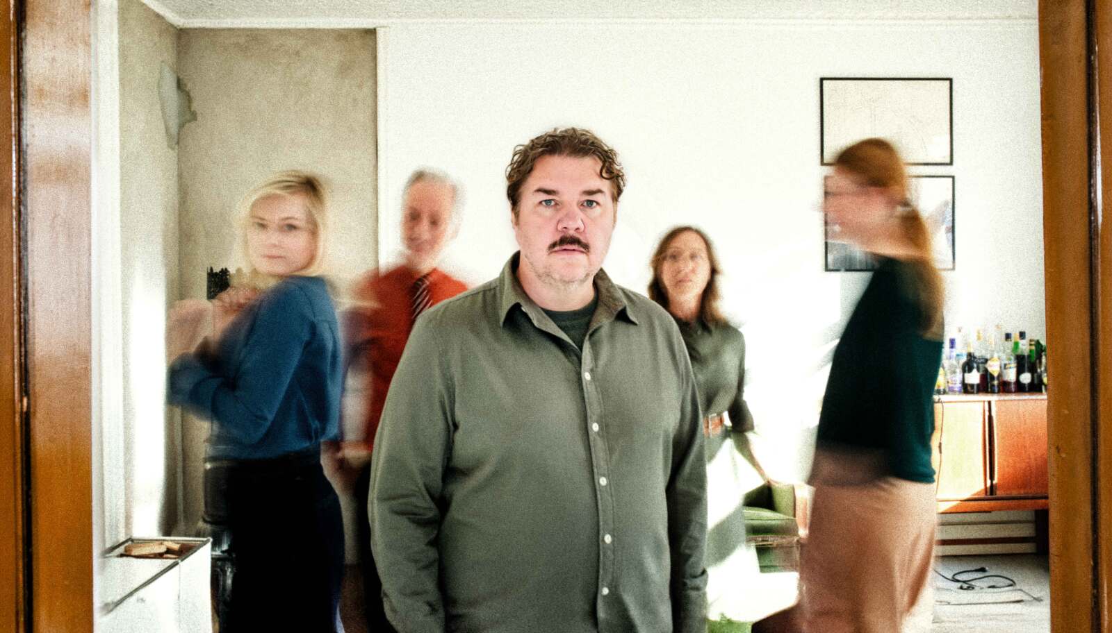 Fem skuespillere i en 60-tallsleilighet. Mannen i midten ser i kamera og er den eneste som ikke er blurry