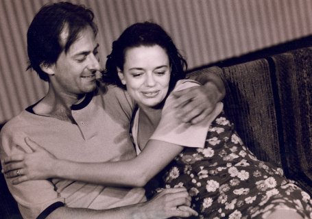 En mann og en kvinne sitter på en sofa, tett omfavnet og smilende; hun har på en blomstrete kjole og han en uformell genser.