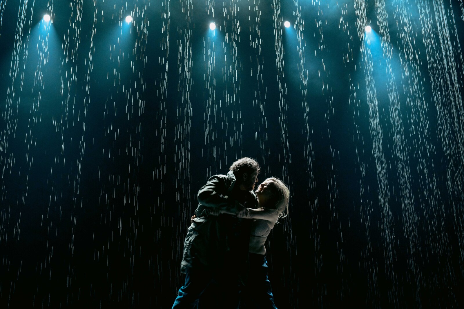 En mann og en kvinne står tett sammen i scenelys mens det regner, i en emosjonell og dramatisk omfavnelse.