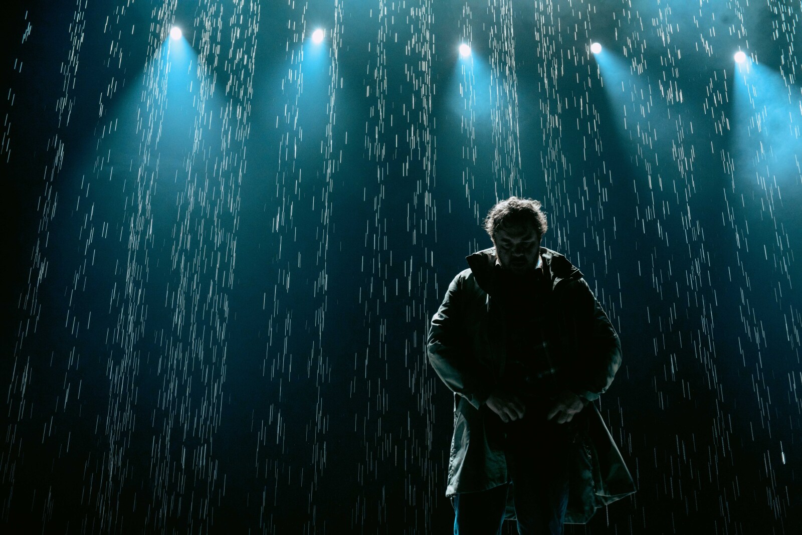 En mann står alene under scenelys og regn, med bøyd hode og alvorlig uttrykk.