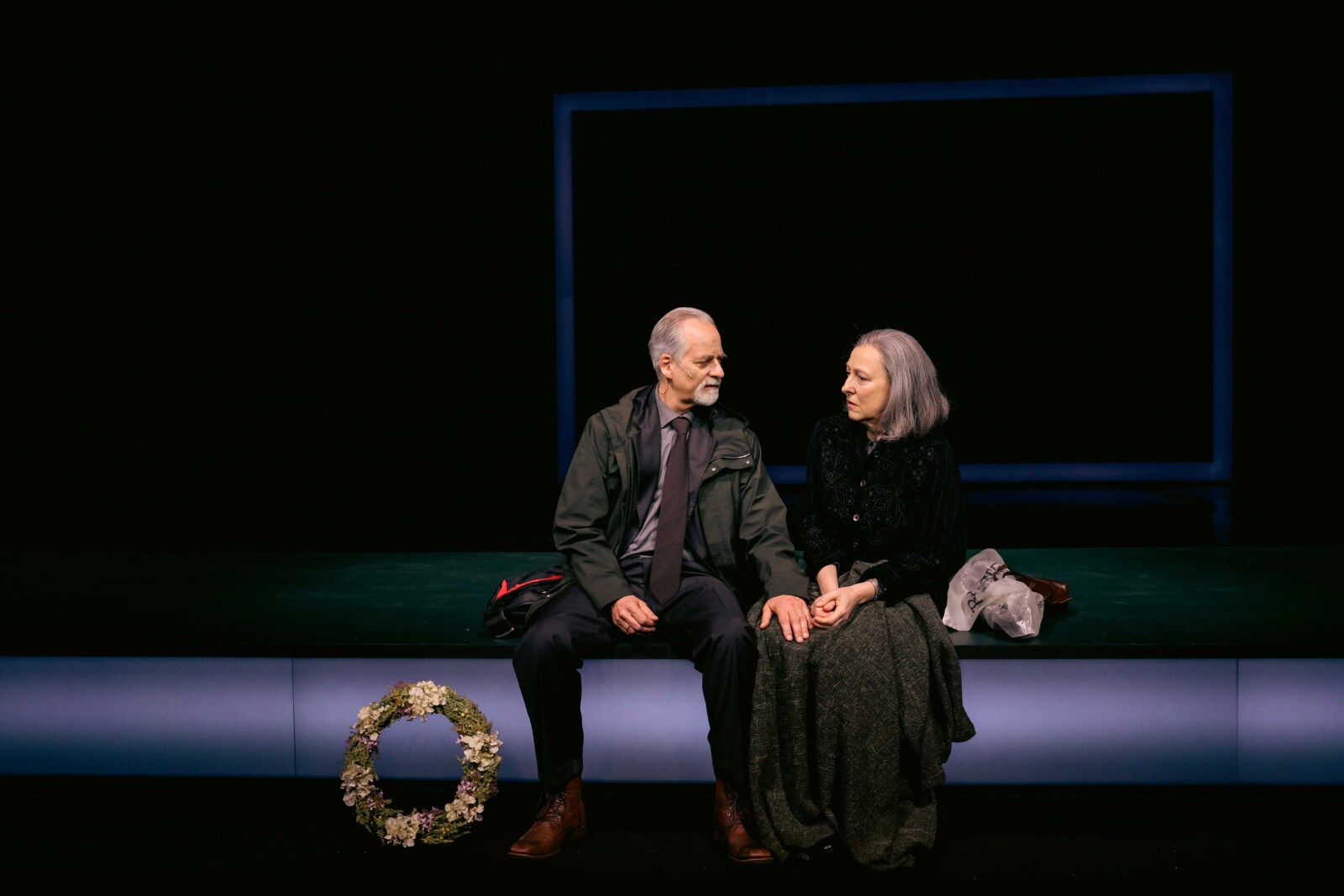 En kvinne og en mann sitter ved siden av hverandre på scenekanten, med en blomsterkrans plassert ved føttene deres. Stemningen virker melankolsk eller ettertenksom.