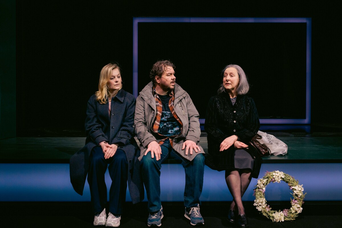 Tre karakterer, to kvinner og én mann, sitter ved siden av hverandre på scenekanten. De ser mot hverandre i samtale, med en blomsterkrans ved den ene kvinnens føtter.