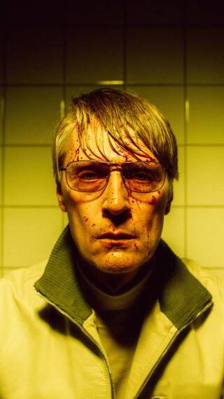 Svett mann med solbriller og blodsprut i fjeset i gult lys. Hvite fliser på veggen bak