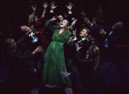 Kvinne synger, mens en gruppe artister står rundt og strekker armene mot henne. Alle er festkledd
