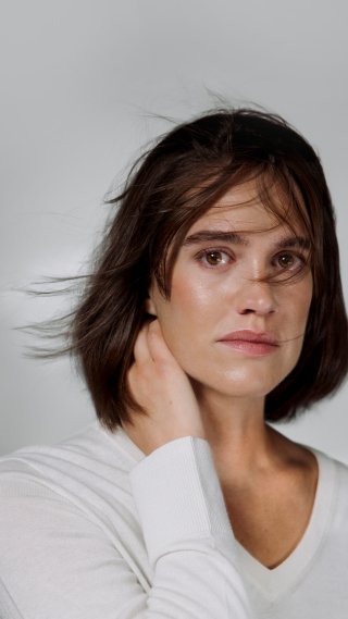 Kvine i hvit genser som holder hånda bak nakken og ser alvorlig i kamera mot hvit bakgrunn