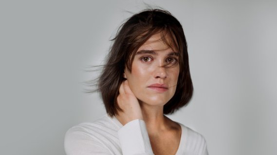 Kvine i hvit genser som holder hånda bak nakken og ser alvorlig i kamera mot hvit bakgrunn