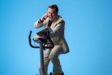 Mann med oppkavet uttrykk på trimsykkel mot blå bakgrunn, med frakk, PC og mobil i hendene, skjorten er delvis åpen og uordnet.