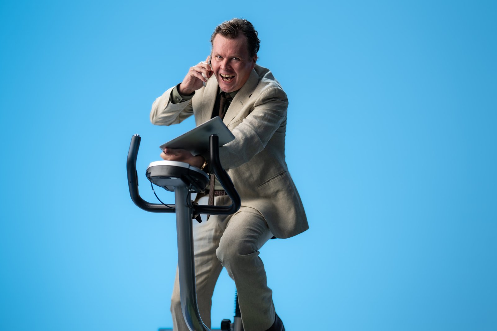 Mann med oppkavet uttrykk på trimsykkel mot blå bakgrunn, med frakk, PC og mobil i hendene, skjorten er delvis åpen og uordnet.