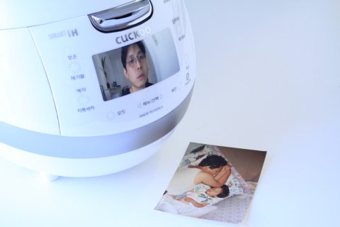 Et bilde av en elektrisk riskoker, hvor det vises bilde av en ung sørkoreansk mann på skjermen. Ved siden av riskokeren ligger det et fotografi av en voksen mann som sover ved siden av et barn.