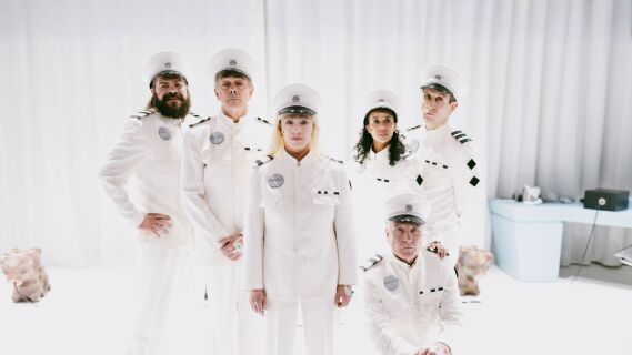 Seks personer i hvite futuristiske kostymer stilt opp til gruppebilde