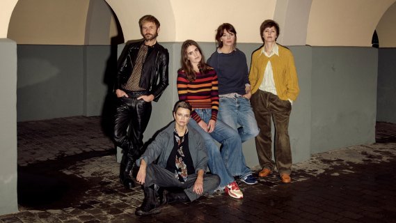 Gruppe med fire kvinner og en mann i en buegang. De er alvorlige, den ene kvinnen sitter, resten står og lener seg mot veggen