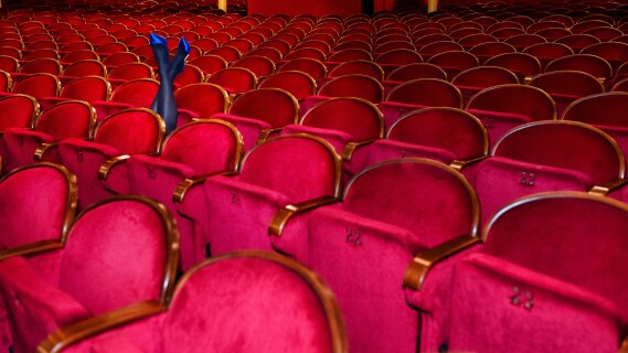 Tom teatersal med røde fløyelsseter og én person som ligger bakover i setet med bena i været, iført strømpebukse og blå høyhælte sko.