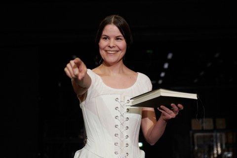 Kvinne i hvit korsett holder bok, smiler og peker fremover