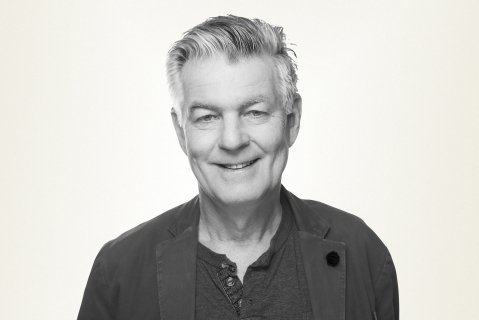 Svart-hvitt-bilde av mann i 70-årene med grått hår og mørk skjorte som smiler til kamera. Lys bakgrunn.